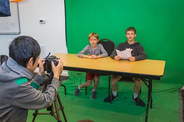 Kids filming a video project at summer camp, seated at a table with a green screen background. Kids filming a video project at summer camp, seated at a table with a green screen background.
