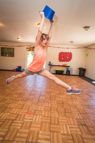 Young camper leaping indoors holding blue hat, showcasing summer camp activities and fun experiences.