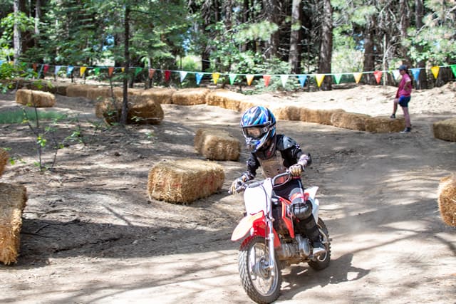 Child riding dirt bike at summer camp, forest trail in background with colorful flags and hay bales marking path. Child riding dirt bike at summer camp, forest trail in background with colorful flags and hay bales marking path.