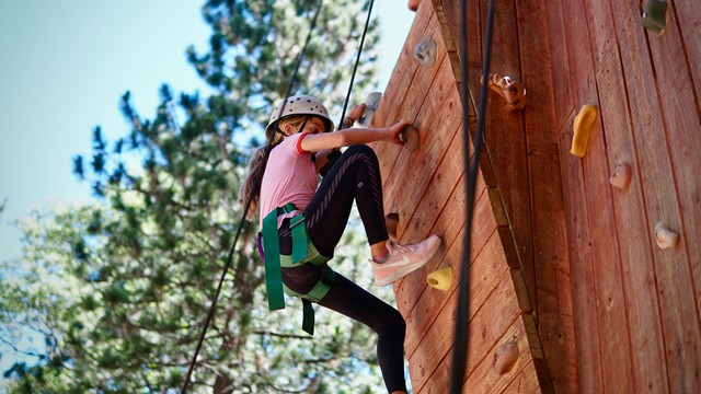 Young girl climbing rock wall at summer camp, wearing safety gear, surrounded by trees. Perfect for adventurous kids. Young girl climbing rock wall at summer camp, wearing safety gear, surrounded by trees. Perfect for adventurous kids.