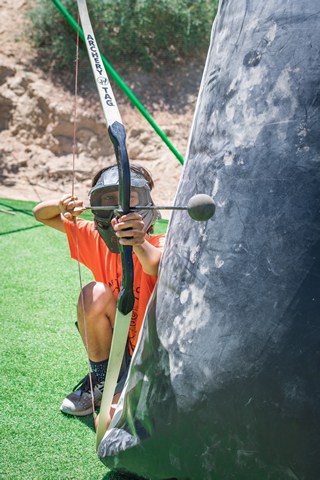Child playing archery tag at summer camp, crouching behind inflatable barrier for fun and safety. Child playing archery tag at summer camp, crouching behind inflatable barrier for fun and safety.