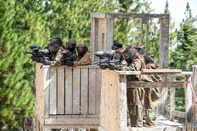 Campers enjoying a paintball game at summer camp, equipped with gear and aiming from a wooden platform amidst trees. Campers enjoying a paintball game at summer camp, equipped with gear and aiming from a wooden platform amidst trees.