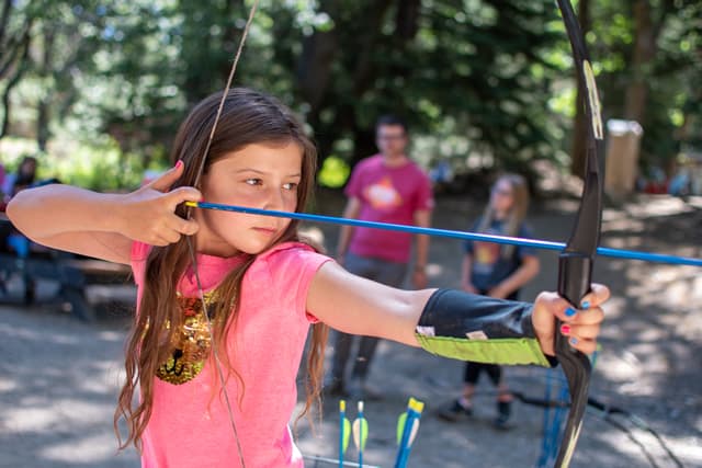 Young girl practicing archery at a summer camp in a forest setting. Young girl practicing archery at a summer camp in a forest setting.