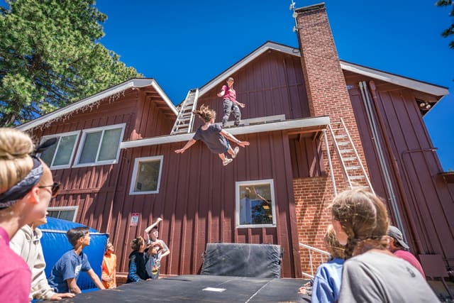 Child mid-air during trampoline activity at summer camp, surrounded by campers and counselors, with rustic cabin backdrop.