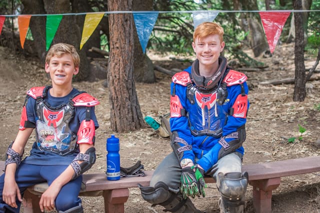 Two campers in motocross gear smiling on a bench at a forest summer camp with colorful flags overhead.