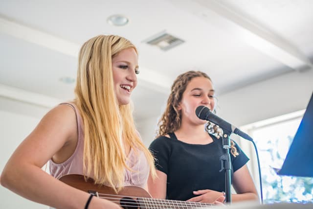 Two girls performing music at a summer camp talent show, one playing guitar and the other singing into a microphone. Two girls performing music at a summer camp talent show, one playing guitar and the other singing into a microphone.