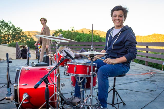 Young camper playing drums outdoors at summer camp talent show. Young camper playing drums outdoors at summer camp talent show.