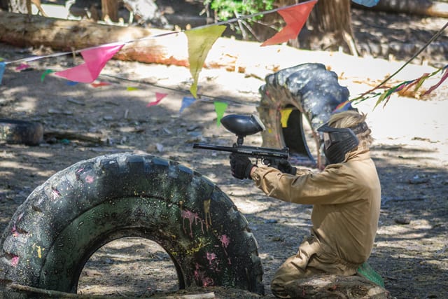 Child playing paintball at a summer camp, aiming behind colorful flags and tires in a forest setting. Child playing paintball at a summer camp, aiming behind colorful flags and tires in a forest setting.