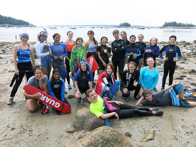 Group of teenagers in wetsuits enjoying a beach day at summer camp, with boats in the background. Group of teenagers in wetsuits enjoying a beach day at summer camp, with boats in the background.