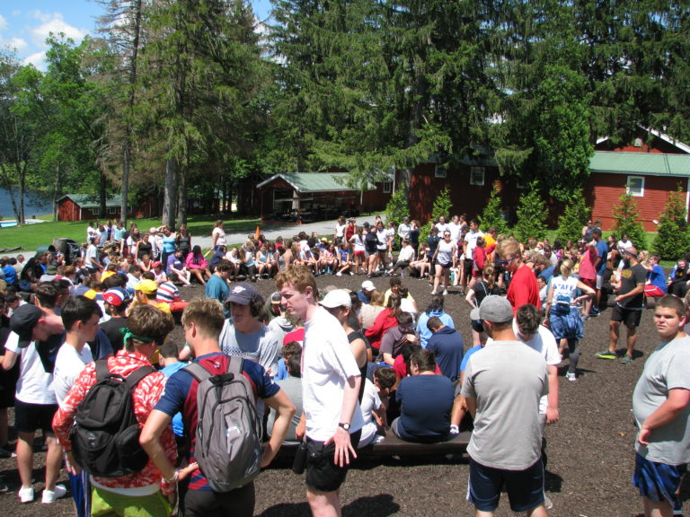 Outdoor summer camp gathering with groups of kids and counselors, surrounded by trees and cabins. Outdoor summer camp gathering with groups of kids and counselors, surrounded by trees and cabins.