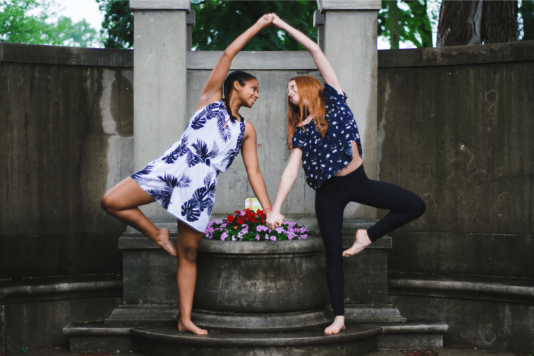 Two friends practicing yoga poses at a summer camp, surrounded by flowers and greenery.