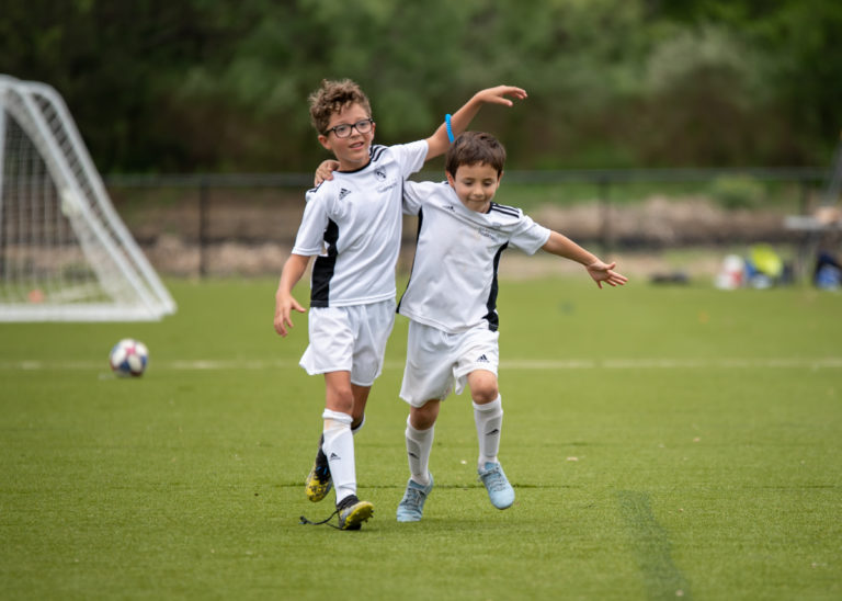 Two kids enjoying soccer at summer camp, running on a green field with a goalpost in the background. Two kids enjoying soccer at summer camp, running on a green field with a goalpost in the background.