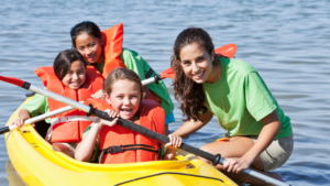 Kids kayaking at summer camp, guided by a counselor in green shirt, smiles and fun on the water. Perfect camp activity!