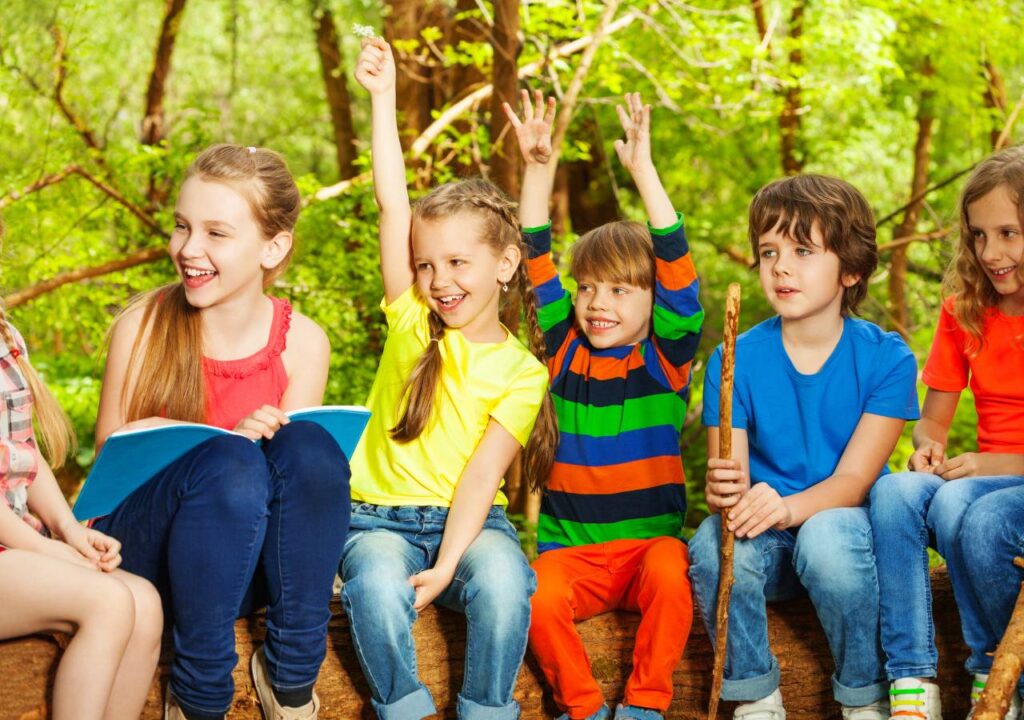 Little boys and girls in different colored clothes smiling and sitting on a log with a background of trees.