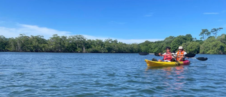 Two campers kayaking on a calm lake surrounded by lush greenery, enjoying outdoor activities at summer camp.
