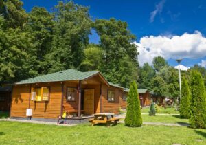 Wooden cabins in a lush summer camp with picnic tables, surrounded by trees under a bright blue sky.