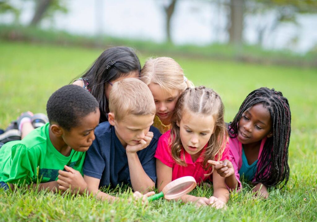 Little boys and girls in different colored clothes smiling and sitting on a log with a background of trees.