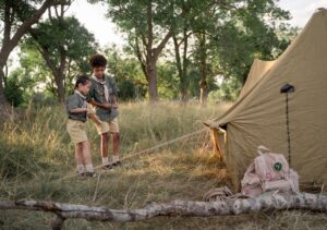 Kids setting up a tent at summer camp, surrounded by trees and grassy field. Perfect for outdoor adventure seekers.