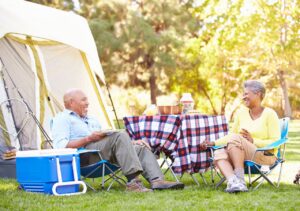 Elderly couple enjoying a summer camp picnic outside a tent, laughing and relaxing at a vibrant campsite.