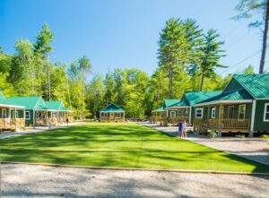 Summer camp cabins with green roofs surrounded by lush trees and a central grassy area for kids' activities.