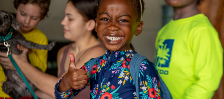 Smiling kid at summer camp giving a thumbs up, surrounded by friends and a dog. Fun and friendship at summer camp!