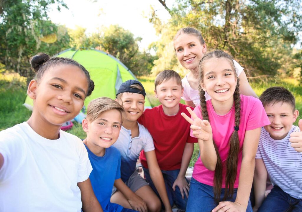 A group of children sitting in front of a tent on a grassy area with smiling faces. They are in a park with trees in the background.