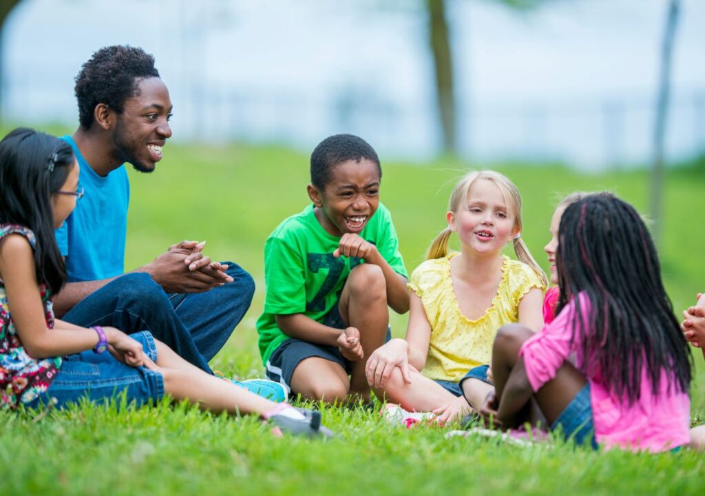 Kids and an adult sitting on the green grass and laughing on summer camp jokes
