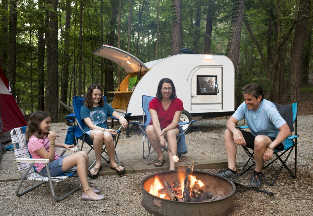 Family enjoying campfire at summer camp with trailer in woods, perfect for relaxing and outdoor activities.