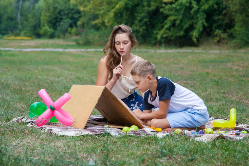 Children crafting at a summer camp, sitting on grass with colorful balloons and art supplies.