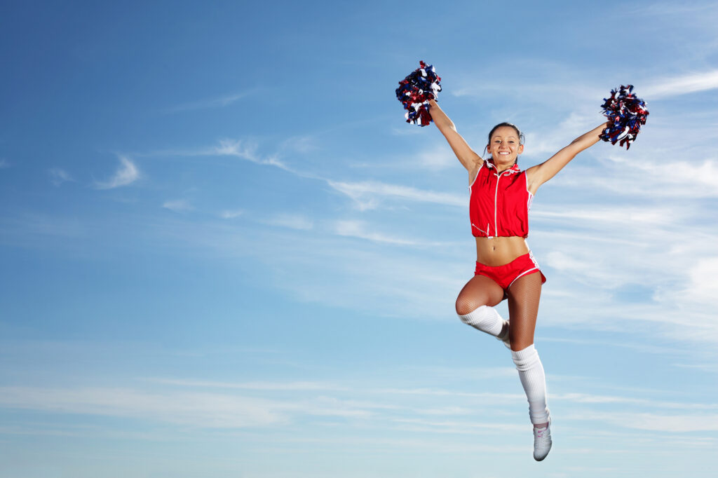 Cheerleader jumping with pom-poms under blue sky at summer camp.