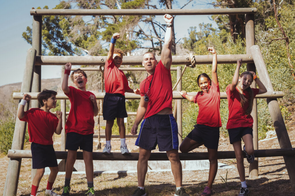 Kids enjoying a summer camp obstacle course with a coach, all in red shirts, surrounded by nature and trees.