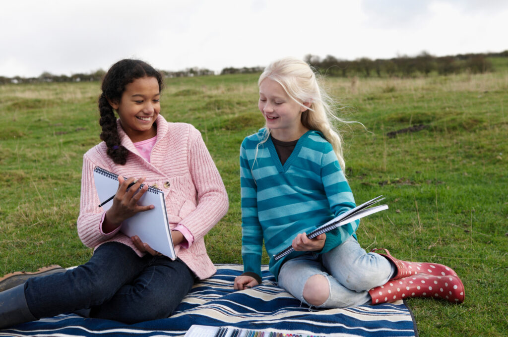 Two girls enjoying outdoor activities and drawing at a summer camp, sitting on a blanket in a grassy field.