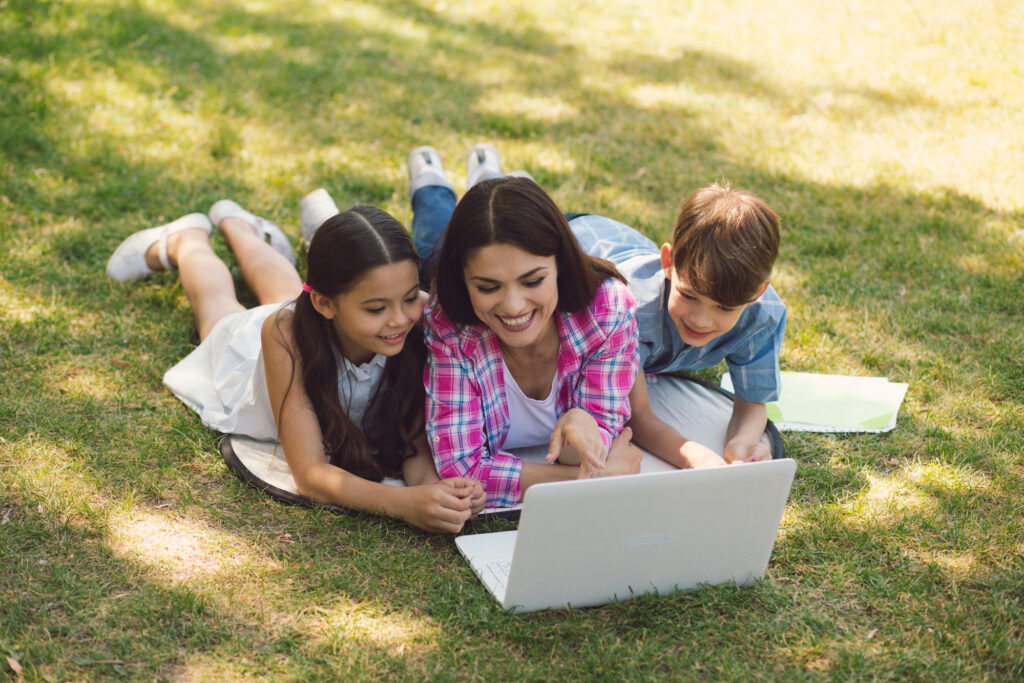 Mom with kids exploring summer camps online, lying on grass and using a laptop, summer camp directory search.