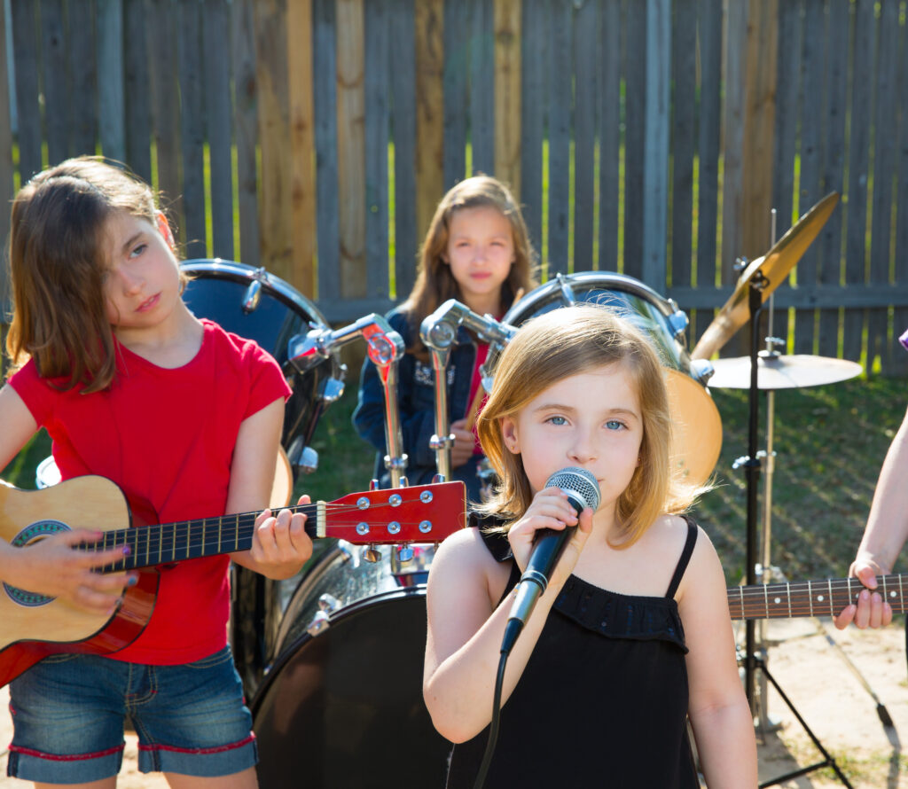 Children playing musical instruments at a summer camp, featuring guitar and drums in an outdoor setting.