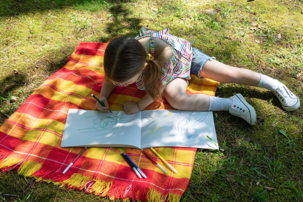 Child drawing outdoors on a picnic blanket at summer camp, enhancing creativity and enjoying nature.