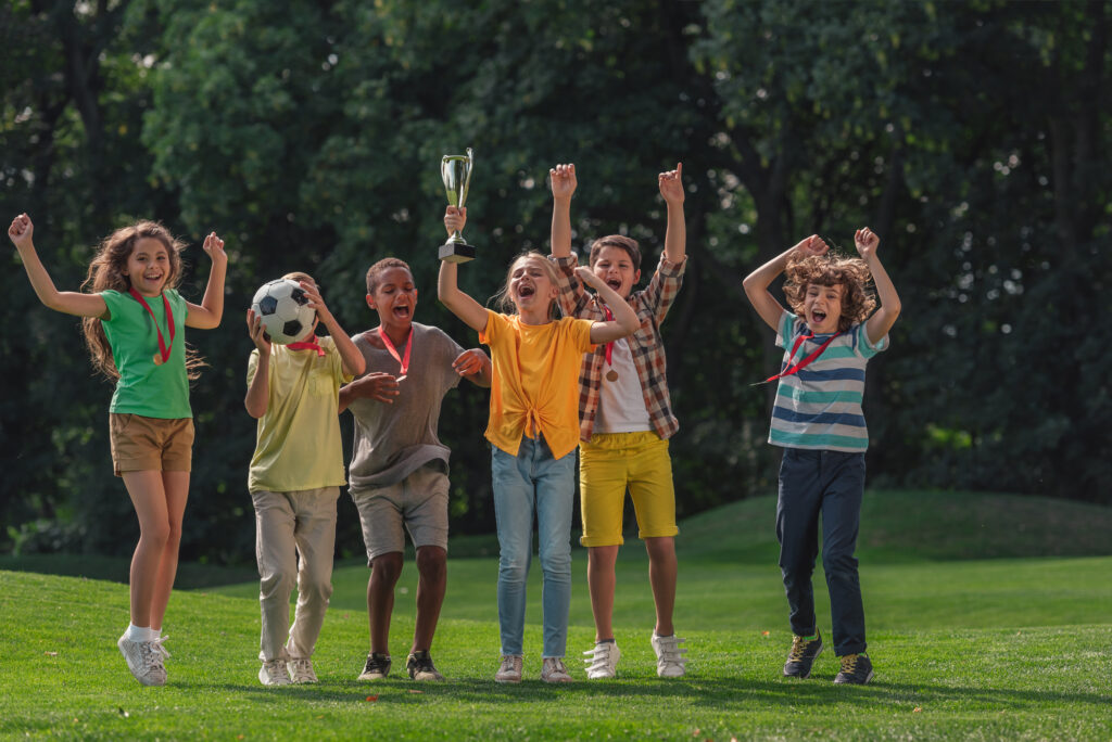 Children celebrating with medals and a trophy at summer camp on a sunny day.