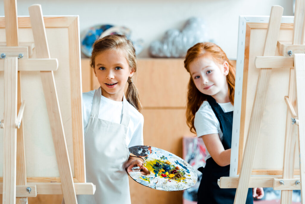 Two kids painting at summer camp art class, wearing aprons and smiling behind easels. Fun and creativity for children!