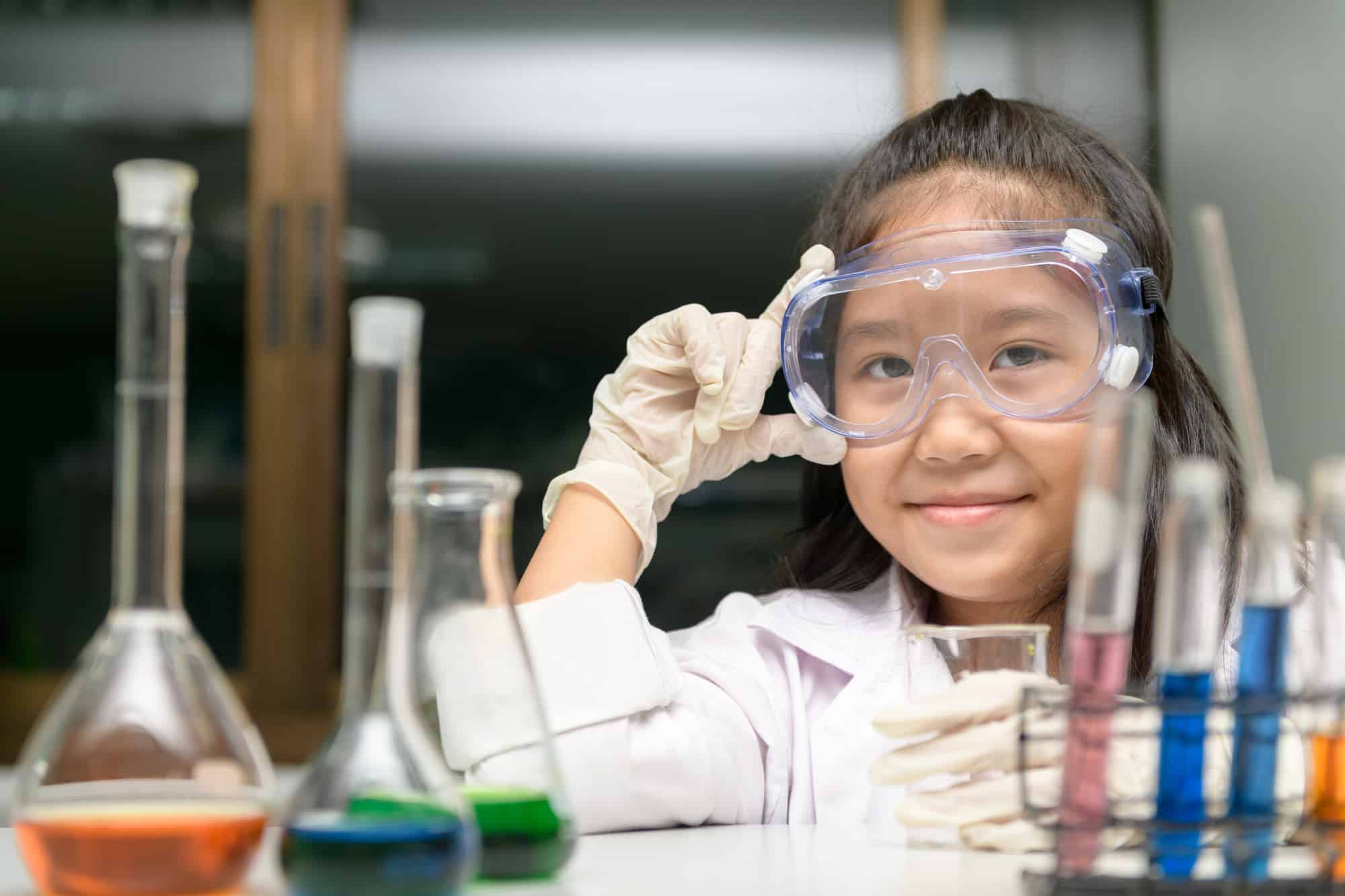 Young girl at science camp wearing goggles, smiling behind colorful test tubes.