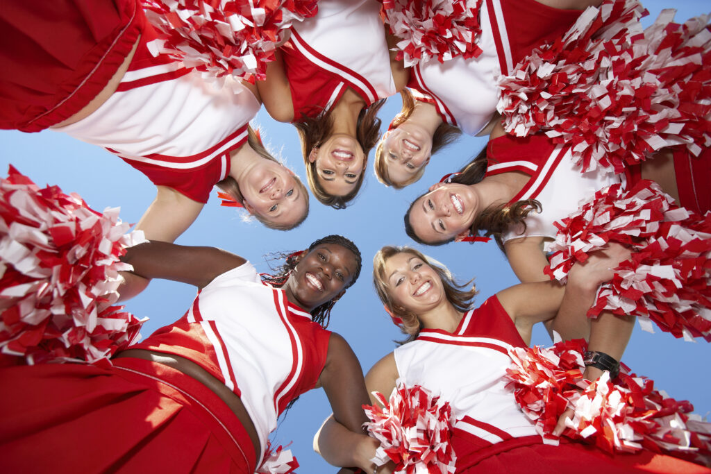 Cheerleaders in red uniforms form a circle, smiling at summer camp under a clear blue sky.