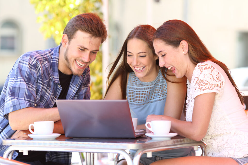Young adults laughing and researching summer camps on a laptop together at an outdoor café.
