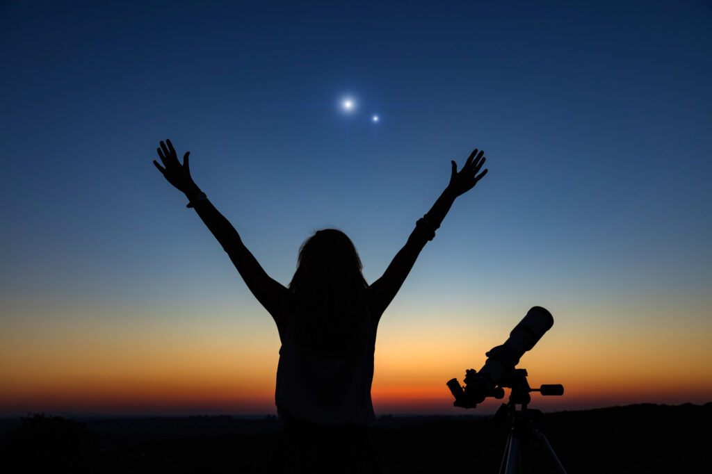 Child stargazing at summer camp with telescope against sunset sky, arms raised in excitement.