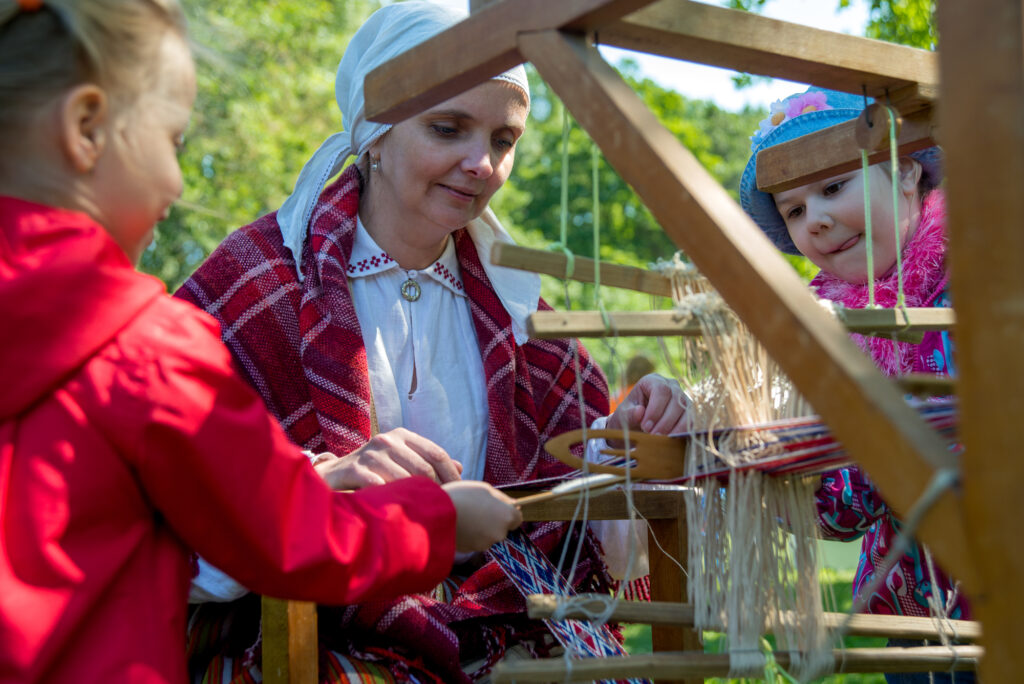 Children learning weaving at a summer camp with guidance from a skilled instructor outdoors.