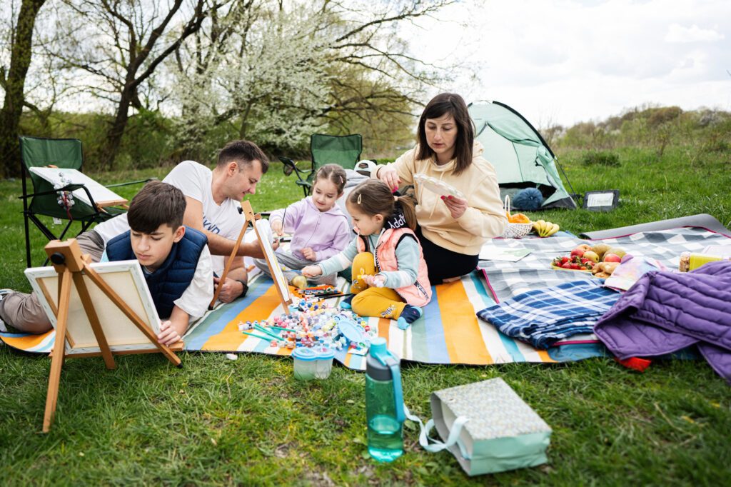 Family enjoying outdoor art activities at a summer camp with picnics and tents in a scenic park setting.