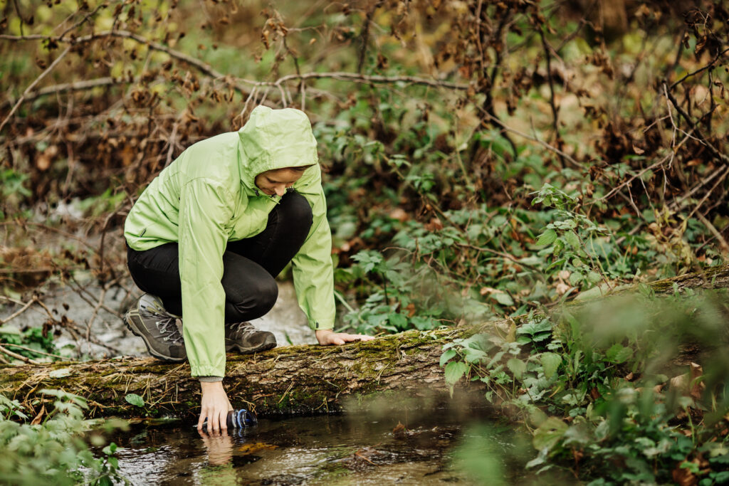 Camper in green jacket collects water in forest creek at summer camp.