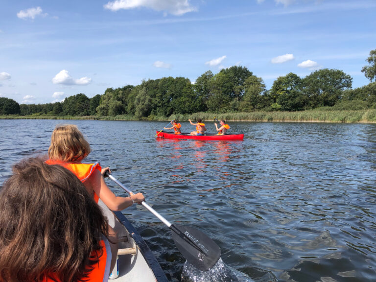 Kids enjoying canoeing on a lake at summer camp, wearing life vests, under a clear blue sky.