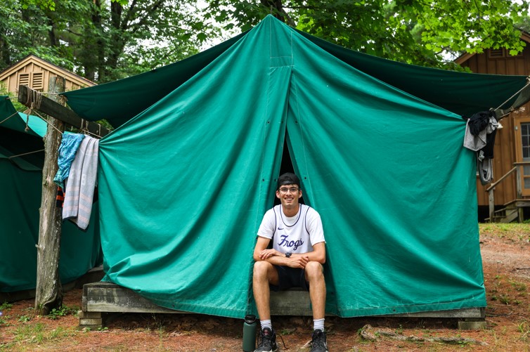 Camper sitting in front of a green tent at summer camp, enjoying the outdoor experience. Camper sitting in front of a green tent at summer camp, enjoying the outdoor experience.