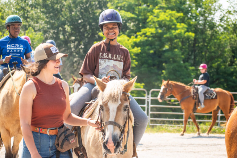Kids enjoying horseback riding at a summer camp with a counselor guiding them outdoors.