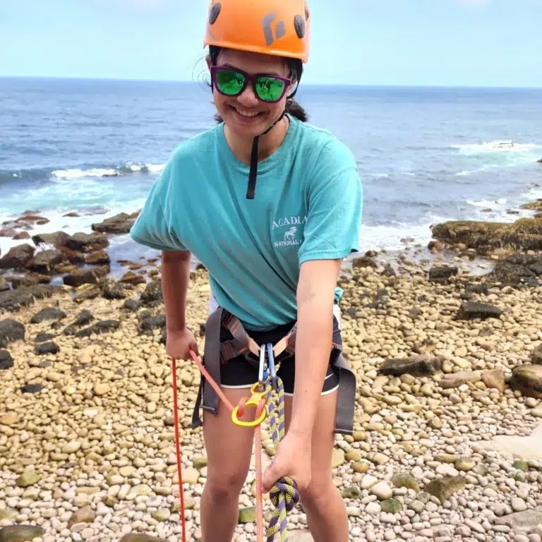 Young camper rock climbing by the ocean at summer camp, wearing safety gear and helmet. Young camper rock climbing by the ocean at summer camp, wearing safety gear and helmet.