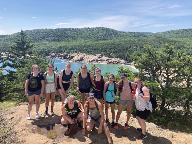 Group of campers hiking with scenic forest and lake view, enjoying summer camp adventures.