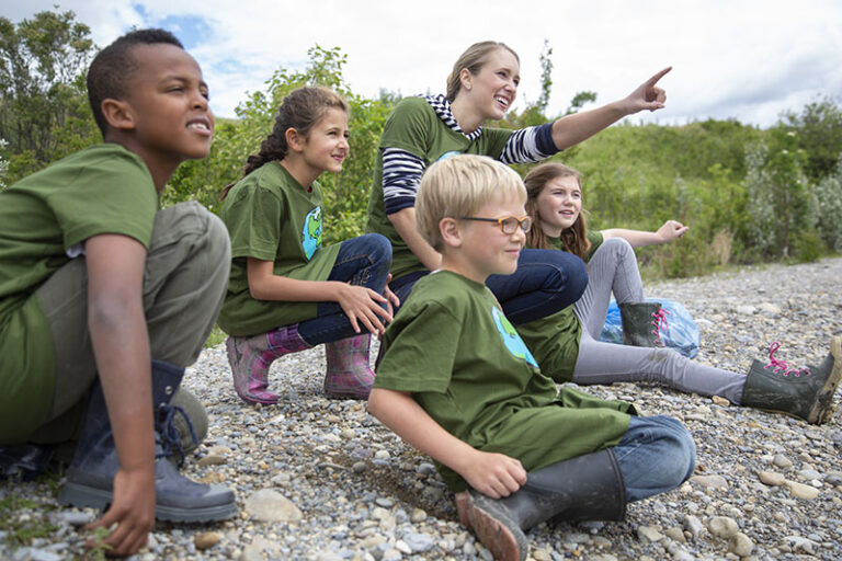 Group of kids exploring nature at summer camp, sitting on rocks with camp leader pointing ahead.